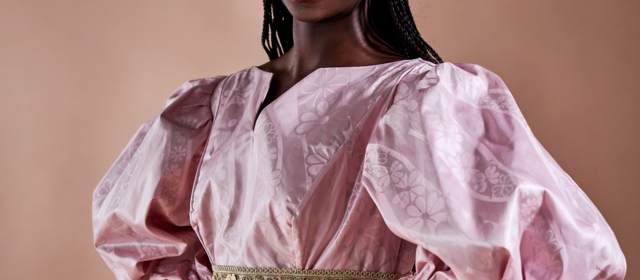 Eine Frau trägt ein zartrosa Damastkleid von Getzner Textil, verziert mit einem detaillierten goldenen Gürtel.