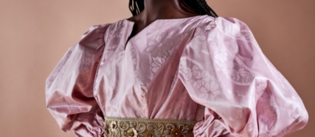Eine Frau trägt ein zartrosa Damastkleid von Getzner Textil, verziert mit einem detaillierten goldenen Gürtel.